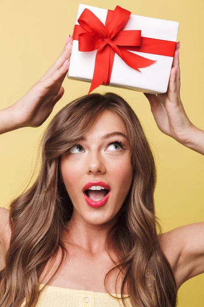 Fotoğraf closeup meraklı kadın gülümseyerek ve mevcut kutusundaki sarı arka plan üzerinde izole başının üstünde yay ile sarılmış tutarak uzun kahverengi saçlı olan heyecanlı