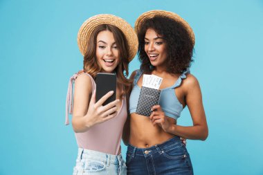 Üzerinde mavi arka plan selfie pasaport ve bilet ile smartphone üzerinde el çekerken gülümseyen iki çok ırklı yaz kız tatil fotoğraf izole