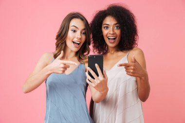 İki neşeli genç kız pembe arka plan üzerinde izole ayakta iken cep telefonu işaret