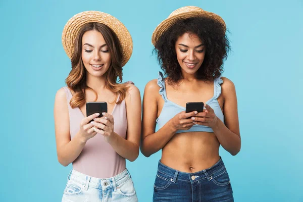 社交的な多民族の女性 代笑顔麦わら帽子との両方を身に着けている青い背景に分離された携帯電話を使用して — ストック写真