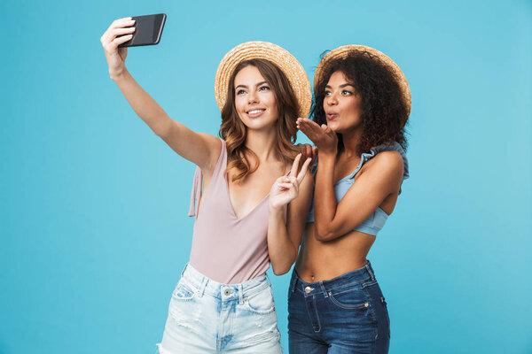 Vacation photo of two multiethnic girls wearing straw hats smiling and showing peace sign at camera, while taking selfie on smartphone isolated over blue background