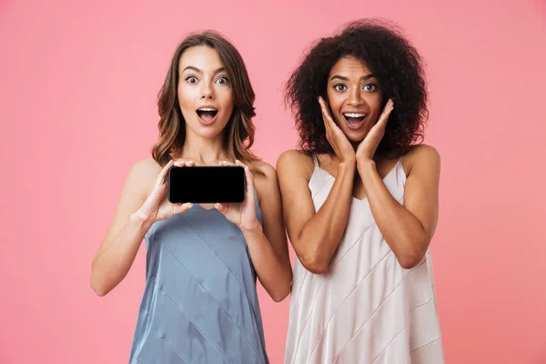 携帯電話のディスプレイを示すピンクの背景に分離立ってかなりショックを受けた若い二人の女性のイメージ — ストック写真