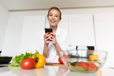 Genç ve güzel kadın alarak resim salataya bir mutfak pişirme sırasında cep telefonuyla