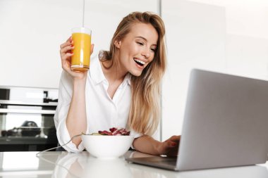 Neşeli genç bir kadın portakal suyu içip kaseden salata yerken bir yandan da mutfakta dizüstü bilgisayardan film izliyor.