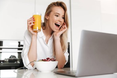 Portakal suyu içme ve süre bir mutfak üzerinde duran ve dizüstü film izlerken bir kase salata yemek genç kadın heyecanlı