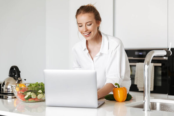 Happy young woman cooking salad while standing on a kitchen with laptop computer