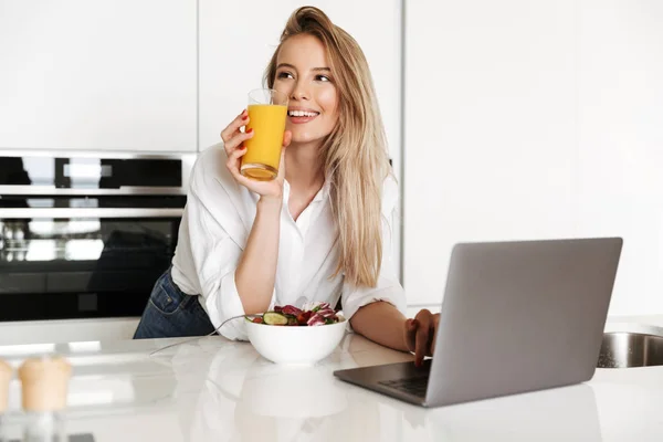 美丽的年轻妇女吃沙拉从碗和喝橙汁 而站在厨房和看电影的笔记本电脑 — 图库照片