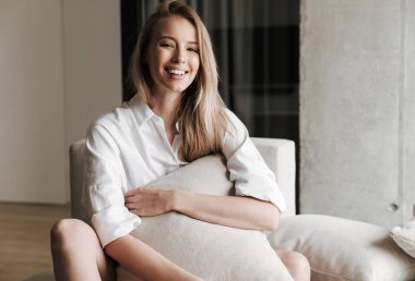 Gülen genç kadın ile bir yastık evde bir kanepede rahatlatıcı ve kameraya bakarak beyaz gömlek giymiş