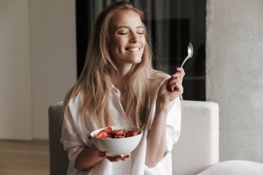 Memnun genç kadın kase meyve mısır gevreği ile evde bir kanepede kapalı gözlerle dinlenirken holding beyaz gömlek giymiş