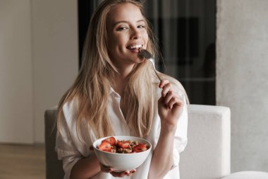 Süre evde bir kanepede rahatlatıcı ve kameraya bakarak kâse Mısır gevreği ile meyve tutan beyaz gömlek giymiş genç kadın gülüyor