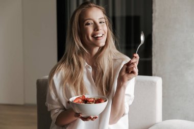 Süre evde bir kanepede rahatlatıcı ve kameraya bakarak kâse Mısır gevreği ile meyve tutan beyaz gömlek giymiş mutlu genç kadın