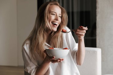 Evde bir kanepede rahatlatıcı ve uzağa arıyorsunuz kâse Mısır gevreği ile meyve tutan beyaz gömlek giymiş neşeli genç kadın