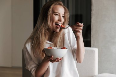 Çekici genç kadın kase meyve mısır gevreği ile evde bir kanepede rahatlatıcı ve uzağa arıyorsunuz holding beyaz gömlek giymiş