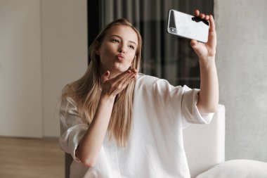 Güzel bayanı selfie cep telefonu ile alarak ve evde bir kanepede otururken öpücük gönderme beyaz gömlek giymiş