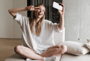 Neşeli genç kadın selfie ile cep telefonu alarak ve evde bir kanepede otururken barış hareketi gösteren beyaz gömlek giymiş