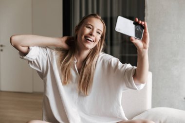 Neşeli genç kadın selfie cep telefonu ile evde bir kanepede otururken alarak beyaz gömlek giymiş