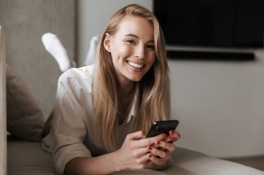 Evde bir kanepede döşenmesi ve kameraya seyir sırasında cep telefonuyla beyaz gömlek giymiş neşeli genç kadın