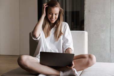 Evde bir kanepede dinlenirken dizüstü bilgisayar holding beyaz gömlek giymiş neşeli genç kadın