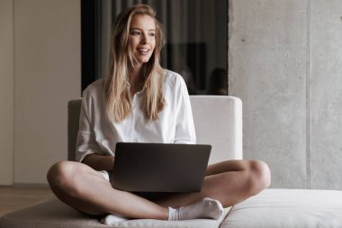 Evde bir kanepede rahatlatıcı ve uzağa arıyorsunuz dizüstü bilgisayar holding beyaz gömlek giymiş genç ve güzel kadın