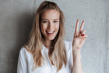 Sevimli güzel genç kadın kapalı kamera gösteren barış hareketi seyir gri duvarın üzerinden görüntü.