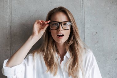 Beyaz gömlek ve gri duvar arka plan üzerinde duran ve kameraya bakarak gözlük çekici genç kadın giyinmiş