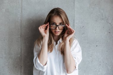 Beyaz gömlek ve gri duvar arka plan üzerinde duran ve kameraya bakarak gözlük giymiş genç ve güzel kadın