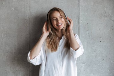 Beyaz gömlek gri duvar arka plan üzerinde duran kulaklık ile müzik dinlemek heyecanlı genç kadın giyinmiş
