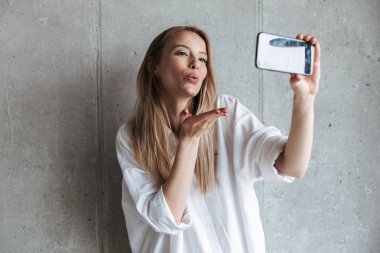 Neşeli bir genç kız portresi bir selfie alarak ve üfleme ayakta gri duvar arka plan üzerinde öp