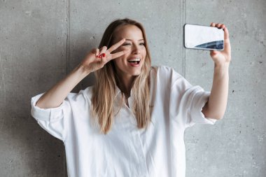 Bir selfie alarak ve barış hareketi duran gri duvar arka plan üzerinde gösterilen mutlu bir genç kız portresi