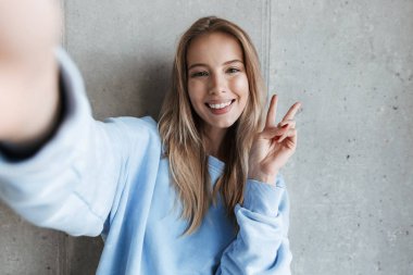 Genç neşeli mutlu güzel kadın tarafından kamera barış hareketi gösteren gri duvar arka plan yapmak selfie üzerinde duran resim.