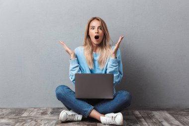 Sürpriz genç sarışın kız bacaklar ile otururken dizüstü bilgisayar kullanan izole gri arka plan üzerinde çarpı işareti