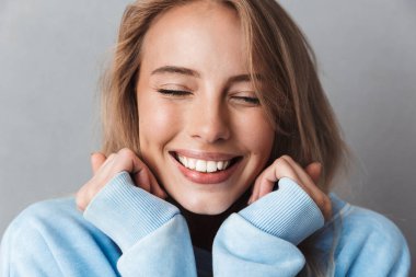 Gri arka plan üzerinde izole gülümseyen genç bir kızın gözleri kapalı ile mavi sweatshirt içinde yakın çekim