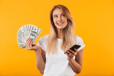 Para banknot demet gösterilen ve cep telefonuyla sarı arka plan üzerinde izole bir gülümseyen dalgın genç sarışın kız portresi