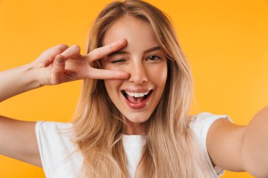 Neşeli genç sarışın kız sarı arka plan üzerinde izole outsretched el ile bir selfie çekerken barış hareketi gösterilen