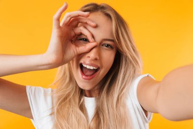 Neşeli genç sarışın kız sarı arka plan üzerinde izole outsretched elleri olan bir selfie çekerken ok jest gösterilen