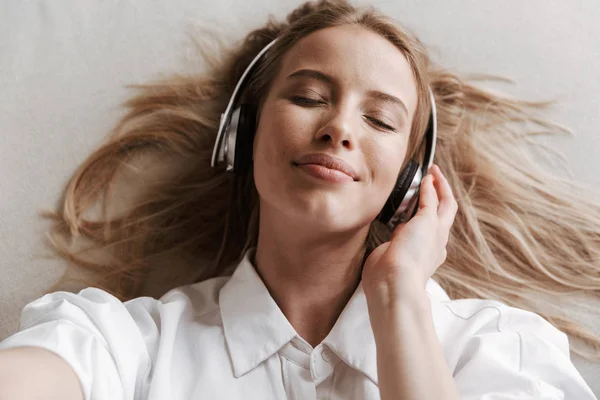 满意的年轻妇女的顶部视图在戴着闭着眼睛的沙发上听音乐时 用耳机自拍 — 图库照片