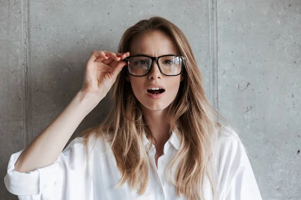 Beyaz gömlek ve gri duvar arka plan üzerinde duran ve kameraya bakarak gözlük çekici genç kadın giyinmiş