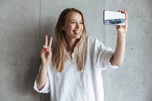 Bir selfie alarak ve barış hareketi duran gri duvar arka plan üzerinde gösterilen neşeli bir genç kız portresi