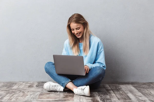 Счастливая Молодая Блондинка Используя Ноутбук Сидя Скрещенными Ногами Сером Фоне — стоковое фото