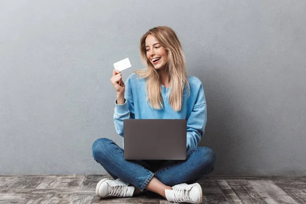 足と一緒に座っている間ノート パソコンを使用して満足している若いブロンドの女の子を渡ったし 灰色の背景上分離されたプラスチック製のクレジット カードを表示 — ストック写真