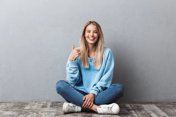 Szczęśliwa Młoda Blondynka Pokazuje Kciuk Górę Gest Siedząc Podłodze Patrząc — Zdjęcie stockowe