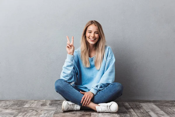 Szczęśliwa Młoda Blondynka Pokazuje Gest Pokoju Siedząc Podłodze Patrząc Kamery — Zdjęcie stockowe