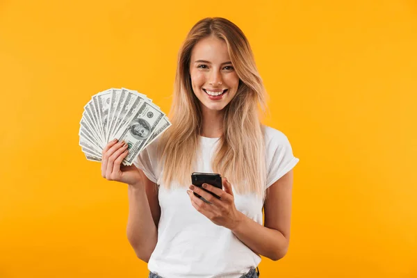 一个快乐的年轻金发女孩的肖像显示一堆钞票和持有手机在黄色背景下隔离 — 图库照片