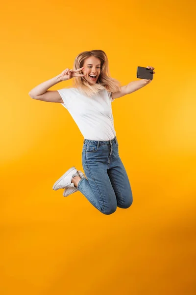 一个快乐的年轻金发女孩的全长肖像显示和平的姿态 而跳跃和采取自拍孤立的黄色背景 — 图库照片