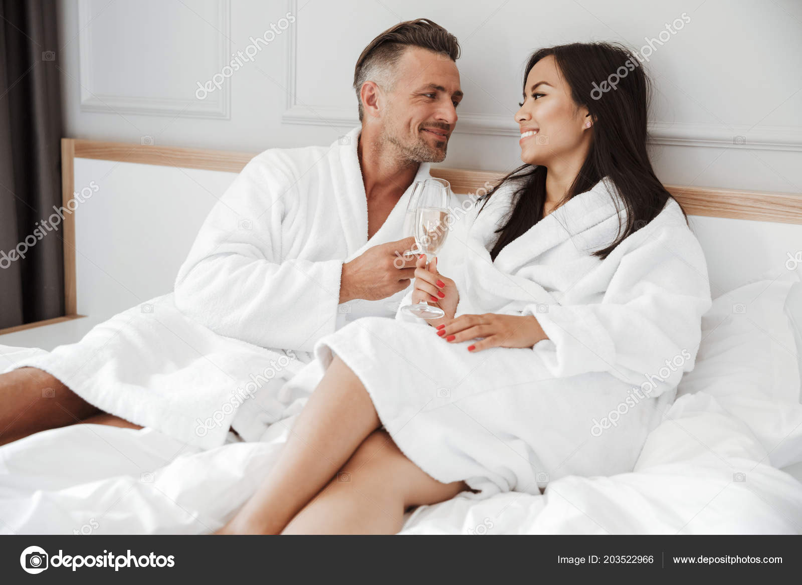 美しいカップル白人男性とアジアの女性がお互いに笑みを浮かべて メガネからスパーク リング ワインを飲みながらホテルの部屋でベッドに横になっている 白いローブを身に着けています ストック写真 C Vadymvdrobot