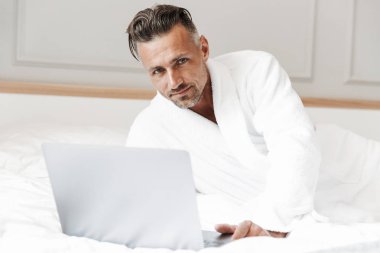 Image of european unshaved man with gray beard wearing white bathrobe looking at camera while lying in bedroom or hotel apartment with silver laptop clipart
