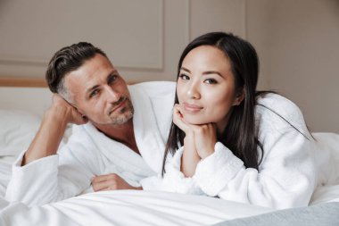 Yatakta yatarken ve kameraya bakarak bornoz giymiş genç çift gülümseyen