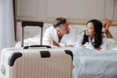 Görüntü bulanık çok ırklı çift erkek ve kadın ön planda büyük bagajlı otel daire yatakta yatan beyaz bornoz giyiyor. Tatil kavramı
