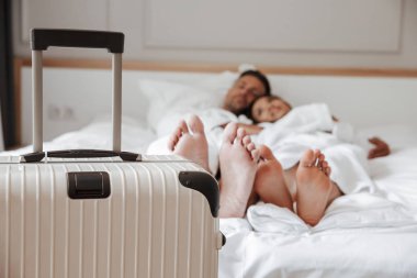 Güzel çift erkek ve kadın yalan ve birlikte ön planda büyük bagaj ile otel odasında yatakta istirahat beyaz bornoz bulanık görüntü. Tatil kavramı