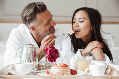 Genç çift gülümseyen kadın besleme adam yatakta yatarken romantik kahvaltı bornoz giymiş.